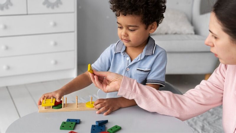 A teacher sitting beside a young child, guiding a wooden shape-sorting activity